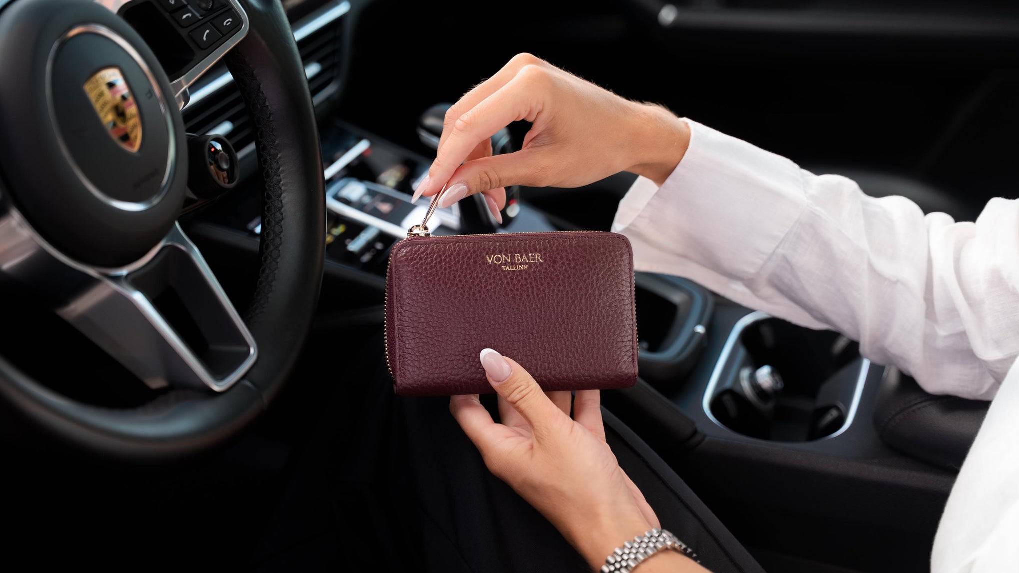 A person in a white shirt holds a beautiful burgundy pebbled leather wallet by Von Baer inside a car, with a Porsche steering wheel visible in the background.