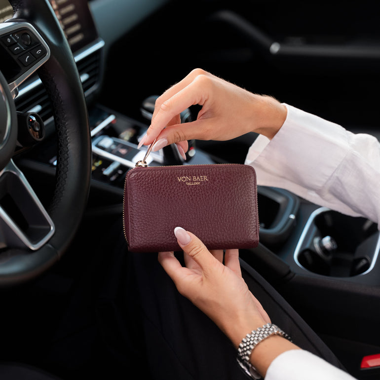 A person in a white dress shirt holds a high quality burgundy pebbled leather wallet with gold hardware inside a black car interior. The wallet features the gold embossed brand name 'VON BAER'.
