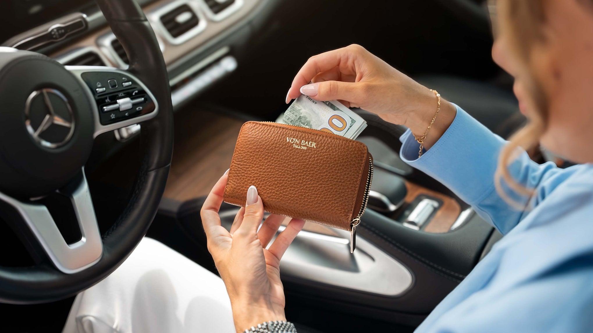 A woman in a blue shirt inserts cash into a high quality brown pebbled leather zip-around wallet with a visible 'Von Baer' logo, held near the car steering wheel inside a mercedes-benz car.
