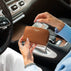 A person sitting in a car holds a brown textured leather wallet with the brand name 'VON BAER' printed on the front, while inserting a dollar bill into it