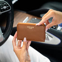 A person holding a classic tan pebbled leather zip-around wallet with gold lettering that reads 'VON BAER' inside a car interior.