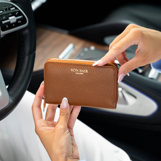 A person holding a classic tan pebbled leather zip-around wallet with gold lettering that reads 'VON BAER' inside a car interior.