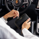 A person inside a car holds a black pebbled leather wallet with gold hardware and the brand name 'Von Baer' printed on the front. The car interior, including a Porsche steering wheel, is visible in the background.