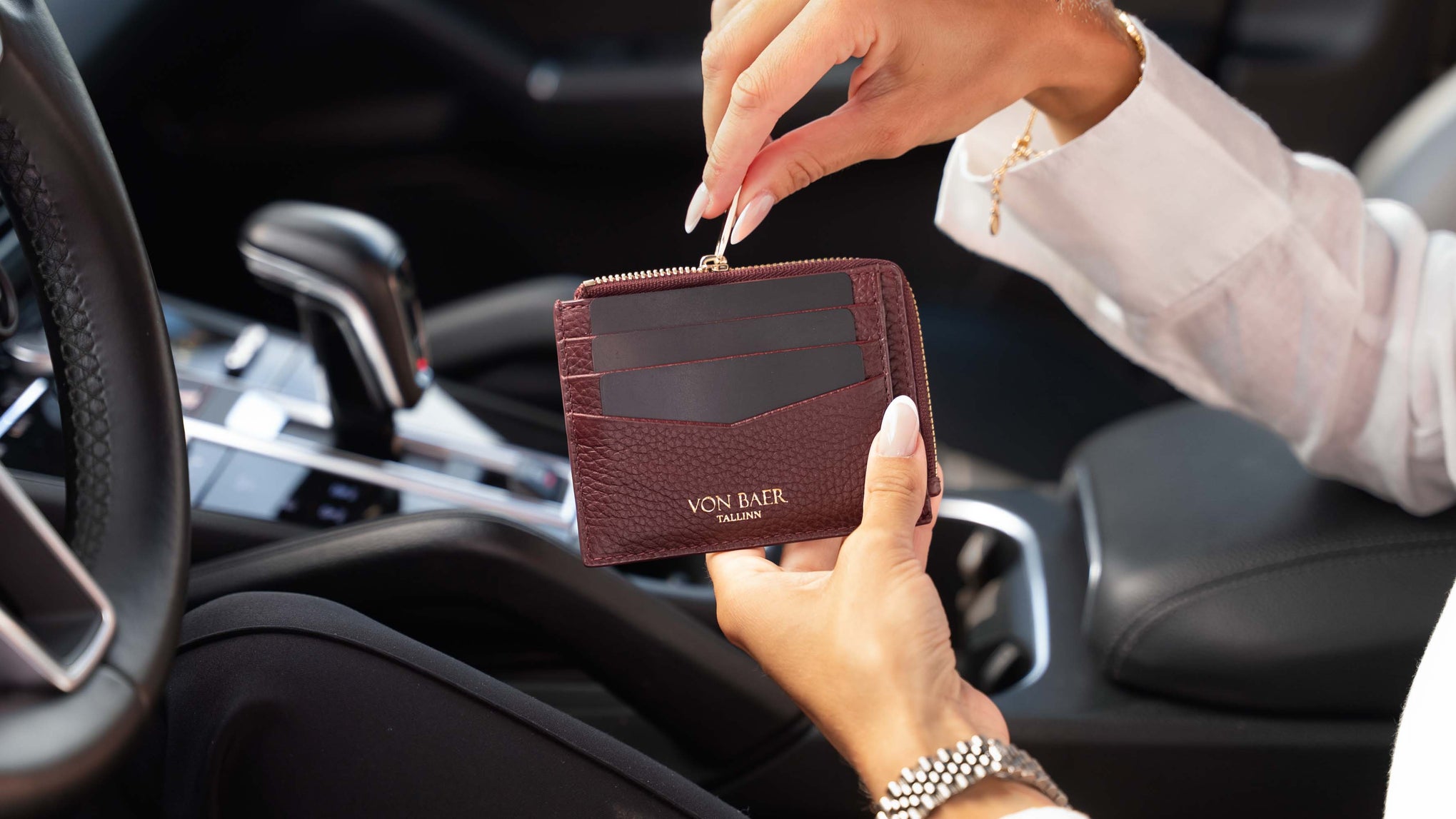 A person holding a small brown luxury leather card holder wallet inside a car, with the brand name 'VON BAER' visible on the front.