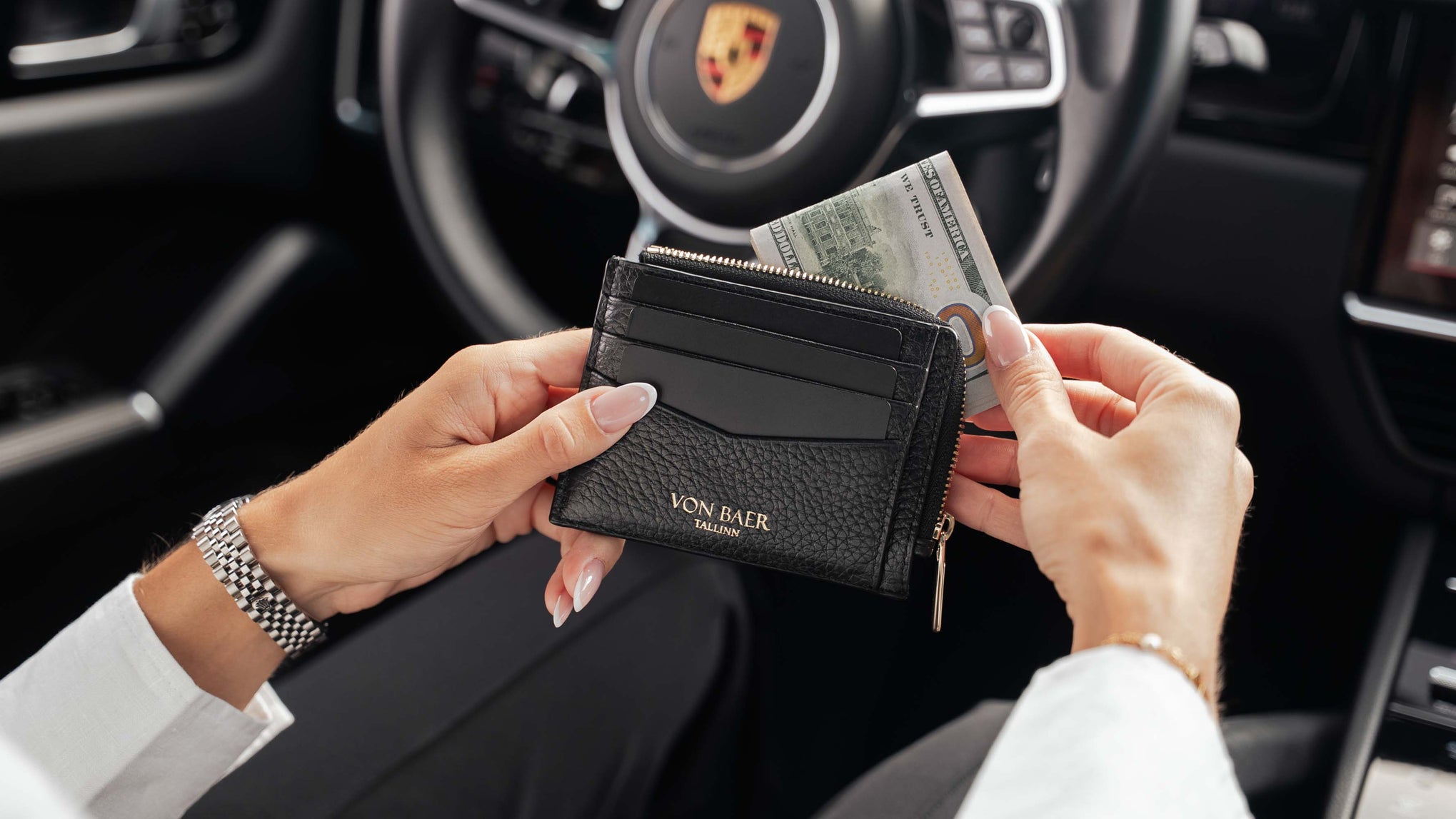 A black pebbled leather wallet with gold lettering reading 'Von Baer Tallinn' is held by a person inside a car, with a US dollar bill partially inserted into the card slots. The Porsche steering wheel is visible in the background.