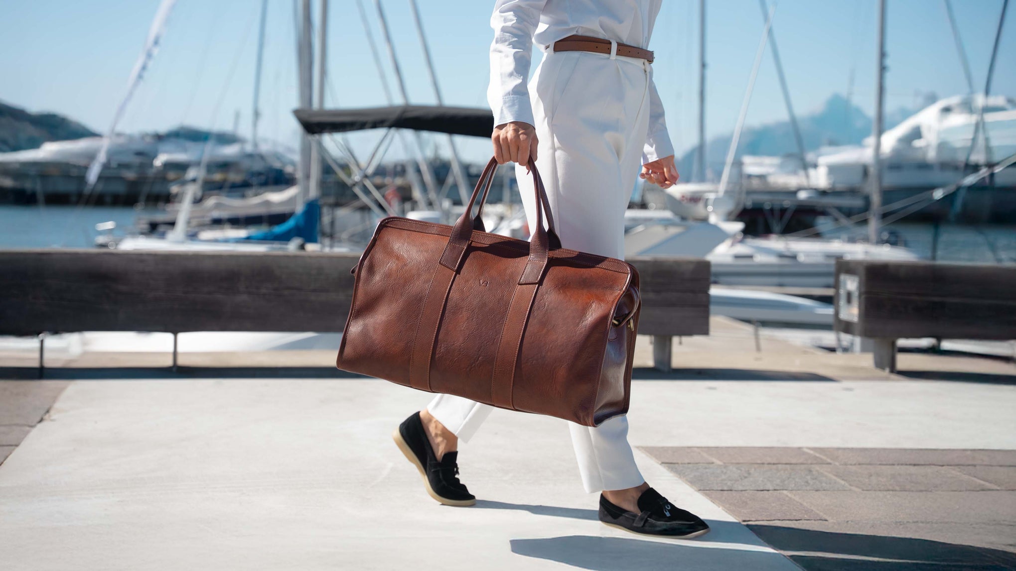 A woman in a white outfit carrying a beautiful solid brown full grain leather duffel bag while walking along a sunny outdoor harbor path with boats, mountains and water in the background.